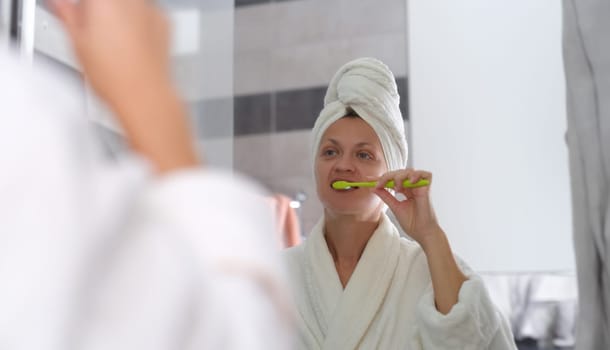 Young woman in bathrobe and with towel on head and brushing teeth in front of mirror in bathroom. Daily hygienic oral care concept