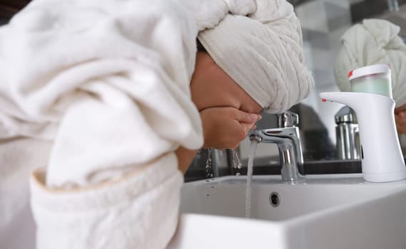 Young woman with towel on head washing face under tap in bathroom. Skin care concept