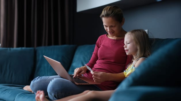 Mother and little daughter are sitting on couch with laptop and credit card. Shopping in online store concept