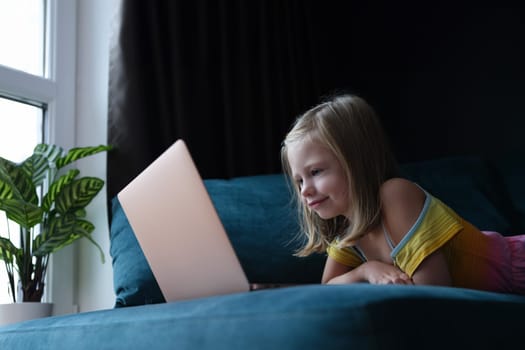 Little girl lying on couch and looking at laptop screen. Homeschooling concept