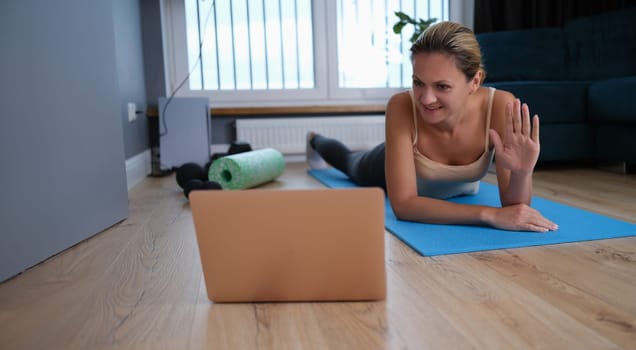 Young woman lying on rubber mat and waving her hand at laptop screen. Remote home fitness concept