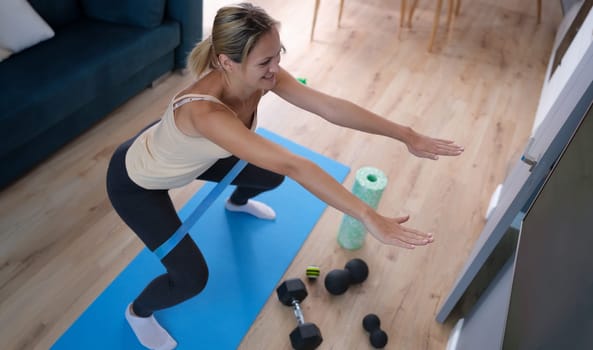 Young woman squatting with elastic band on her legs at home. Remote fitness concept