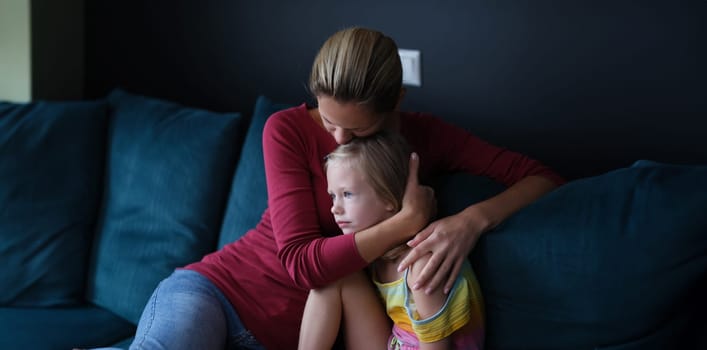 Woman and sad little daughter are sitting on couch and hugging. Maternal support for children concept