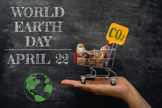 A hand holding a shopping cart with a sign that says CO2 on it. The cart is on a chalkboard with the words World Earth Day April 22