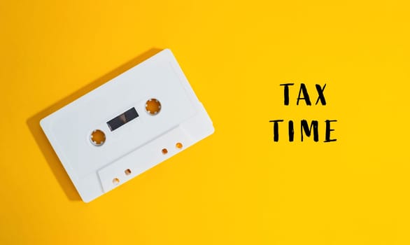 A white cassette tape is on a yellow background with the words Tax Time written below it. Concept of nostalgia and the idea of paying taxes, which can be a tedious and time-consuming task