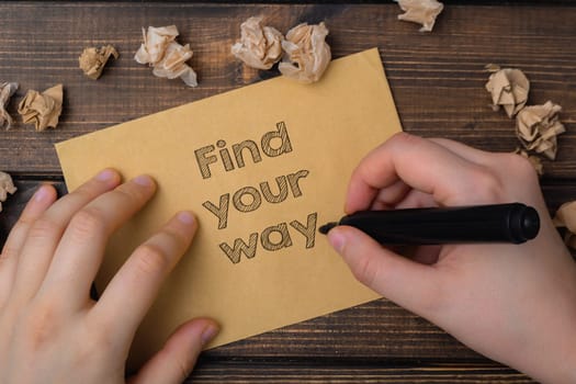 A hand is writing on a piece of paper that says find your way. The paper is on a wooden surface and there are some shredded pieces of paper nearby