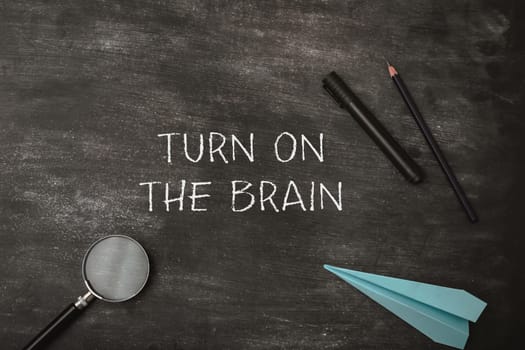 Turn on the brain is written on the chalkboard. The chalkboard is covered with chalk and a pair of pencils. A blue paper airplane is also on the chalkboard