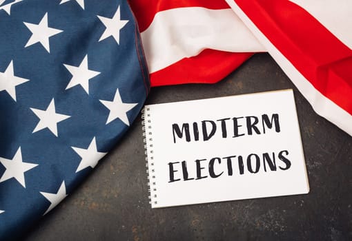 A white piece of paper with the words Midterm Elections written on it. The paper is placed on top of a red and white American flag