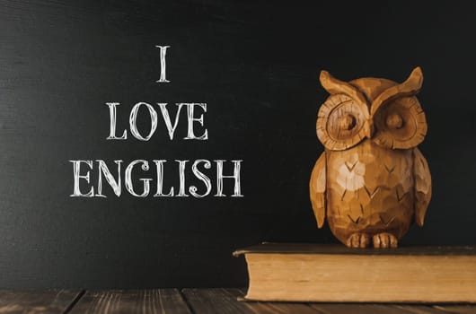A wooden owl sits on a book with the words I love English written on a chalkboard behind it