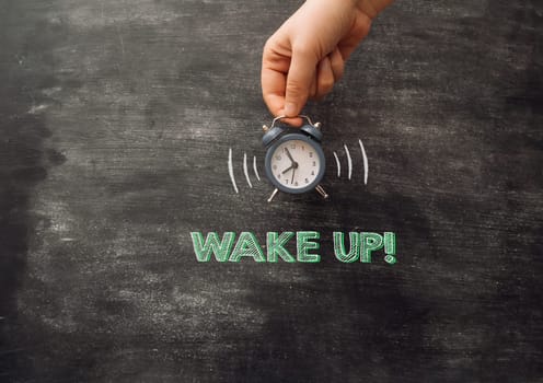 A hand holding an alarm clock with the words wake up written on the chalkboard. Concept of waking up early and the importance of starting the day on time