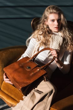 beautiful curly blond hair woman posing with a small shopper brown bag in a vintage chair. Model wearing stylish white sweater and classic trousers