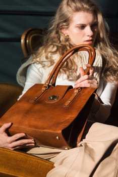 beautiful curly blond hair woman posing with a small shopper brown bag in a vintage chair. Model wearing stylish white sweater and classic trousers