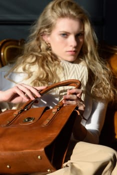 beautiful curly blond hair woman posing with a small shopper brown bag in a vintage chair. Model wearing stylish white sweater and classic trousers