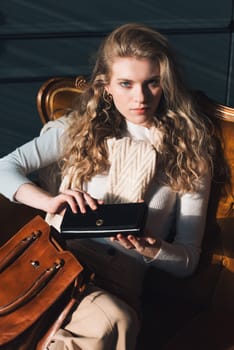 beautiful curly blond hair woman posing with a small shopper brown bag in a vintage chair. Model wearing stylish white sweater and classic trousers