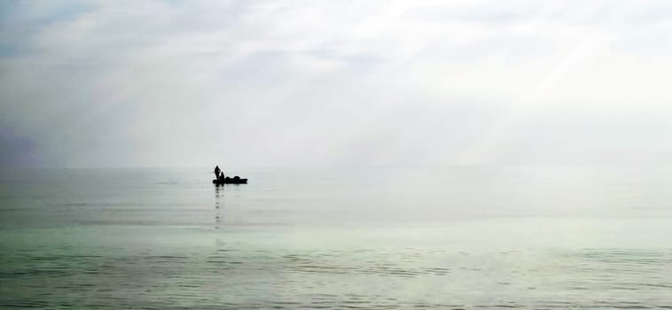Shot of a man on a small catamaran boat by the horizon, which is hard to detect because of the fog and humidity.