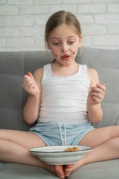 A cute little girl covered in chocolate eats cookies while sitting on the sofa. Vertical photo