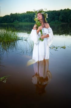 Adult mature brunette woman in a white dress, sundress and a wreath of flowers in summer in water of river or lake in evening at sunset. Celebration of the Slavic pagan holiday of Ivan Kupala