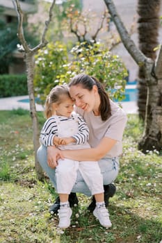 Smiling mother touching head to little girl head on her lap while squatting in garden. High quality photo