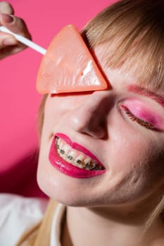 Portrait of a young woman with braces and bright makeup eating a lollipop on a pink background. Vertical photo