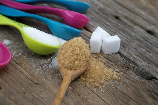 Brown cane sugar and white sugar in colorful plastic measuring spoons spilled around on a rustic wooden surface, measuring utensils in a kitchen setting