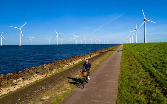 A man is peacefully cycling down a serene path next to a body of water in Flevoland, Netherlands. The scene is surrounded by windmill turbines, creating a picturesque and idyllic setting for a leisurely bike ride.