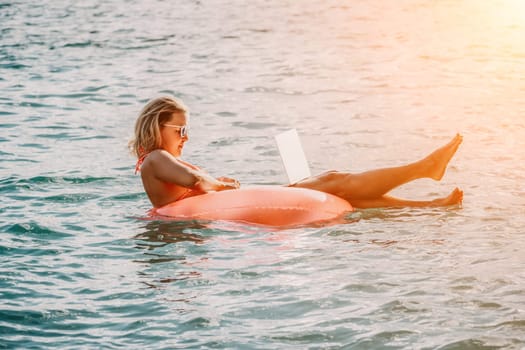 Woman works on laptop in sea. Freelancer, young blond woman in sunglases floating on an inflatable big pink donut with a laptop in the sea at sunset. Freelance, travel and holidays concept