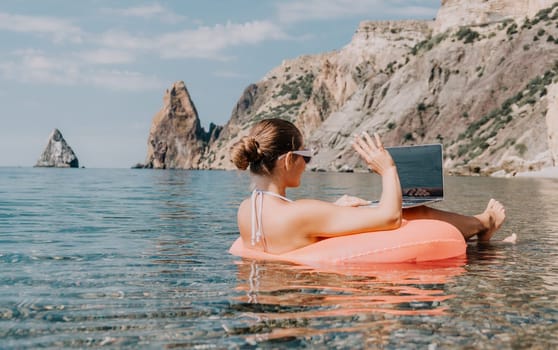 Woman freelancer works on laptop swimming in sea on pink inflatable ring. Pretty lady typing on computer while floating in the sea on inflatable donut at sunset. Freelance, remote work on vacation