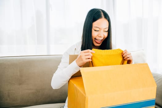 Asian woman on couch opens online package delighted with delivered dress. Smiling and holding the shirt showcasing satisfaction with the purchase. Satisfied buyer concept.