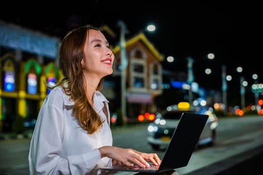 On a bustling city street at night, a woman is hard at work on her laptop computer, utilizing the convenience and mobility of her device to stay connected in the digital age.