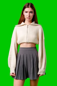 A young woman stands with her hands on her hips against a vivid green backdrop, wearing a fashionable, off-white cropped jacket with a zip and a charcoal gray pleated skirt. Her expression is confident and slightly quizzical, as if challenging the viewer, while her long brown hair frames her face.