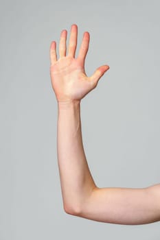 A persons arm is extended upwards with the hand open, showing the palm and fingers clearly against a plain, neutral grey backdrop. The gesture might indicate a question, a desire to speak, or a symbol for stopping.