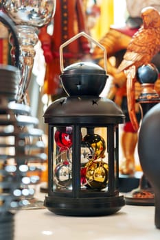 A black, star-patterned lantern filled with shiny red and gold Christmas baubles sits prominently among other holiday decorations. The soft focus background features various ornaments and festive items, creating a warm, seasonal shopping environment.