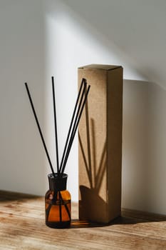An amber glass bottle serves as a simple yet elegant reed diffuser, casting intricate shadows alongside the cardboard packaging on a warm wooden surface bathed in the soft glow of morning sunlight.
