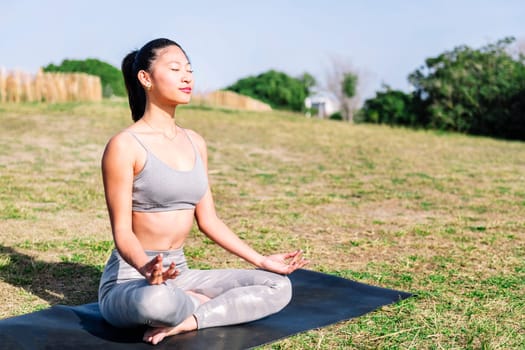 young asian woman doing meditation at park sitting on a yoga mat, concept of mental relaxation and healthy lifestyle, copy space for text