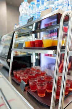 A selection of fruit juice cups, with orange and red hues, are neatly arranged on a metal serving cart, ready for patrons to enjoy in a bustling cafeteria setting. The focus on the front cups, with a blurred background, highlights the refreshment options available.