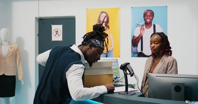 African american deliveryman receiving requested clothes packed in boxes, arriving at fashion showroom to pick up order and sign papers. Woman employee preparing items at cash register. Camera B.