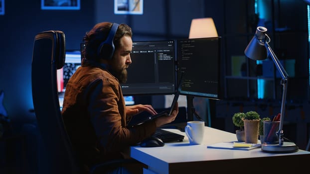 Developer focused on typing complex code in neon lit home office, listening music and developing software application. IT expert wearing headphones, solving coding project tasks on laptop, camera A