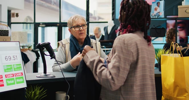 African american employee scanning clothes tags at checkout, preparing bag with products for elderly woman at department store. Retail worker using scanner at cash register, consumerism. Camera B.