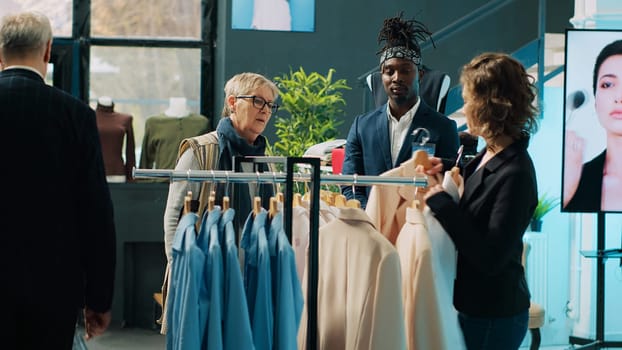 Fashion stylist suggesting clothing items to regular customer, team of store workers help elderly woman to buy trendy clothes on sale. Retail clerks assisting client during shopping spree. Camera A.