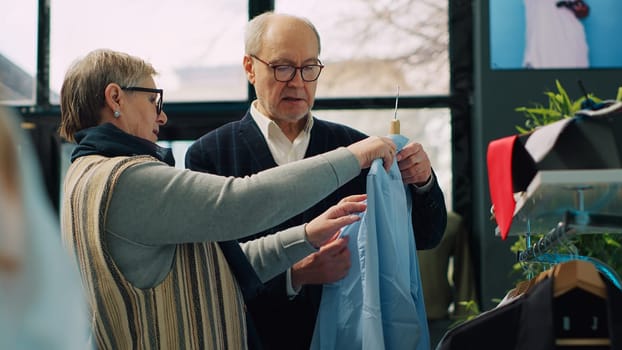 Elderly customers analyzing elegant shirts on hangers at retail store, looking at new clothing line on display at boutique. Senior couple searching for formal wear, posh lifestyle. Camera A.