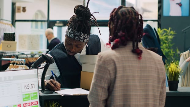 African american deliveryman receiving requested clothes packed in boxes, arriving at fashion showroom to pick up order and sign papers. Woman employee preparing items at cash register. Camera A.