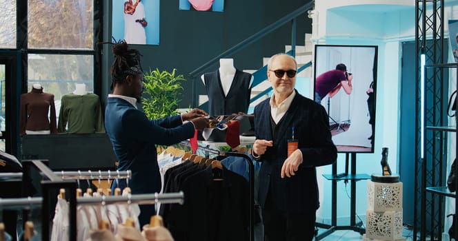 Famous couturier giving order to store assistant to help with finding a tie for his african american client, experienced senior tailor in. Fashion designer shopping for luxury accessories. Camera B.