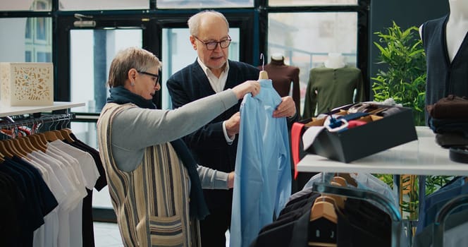 Senior couple shopping for formal outerwear at fashion boutique, looking for trendy elegant clothing items to create new outfits for elderly man. People searching for stylish merchandise. Camera B.