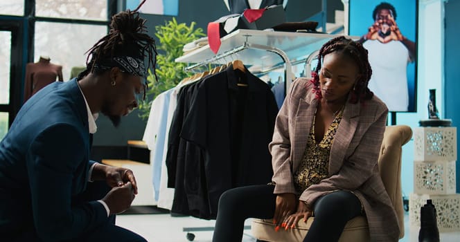 African american store employee helping woman to find the right shoe size, pregnant customer looking to buy comfortable footwear from local shopping center. Client trying on winter boots. Camera B.