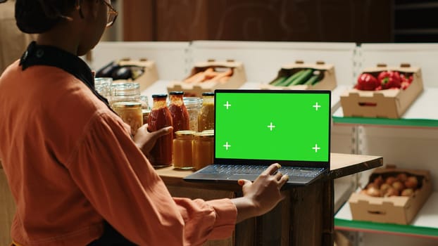 Young vendor looks at greenscreen on laptop while preparing merchandise for customers, using isolated mockup layout on screen. African american seller checking blank chromakey display. Camera 1.