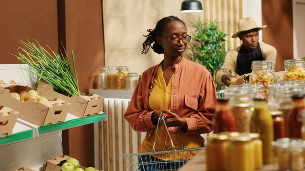 Regular customer searching for eco friendly food alternatives, looking at homemade ethically sourced bulk items at local supermarket. African american vegan woman going grocery shopping. Camera 1.