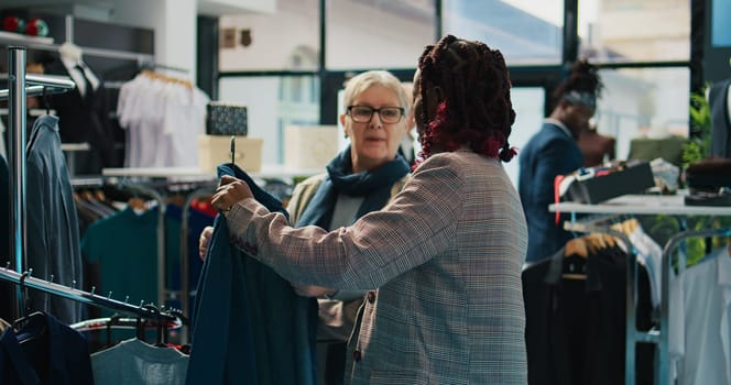 African american retail clerk showing new fashion collection to senior client in department store at the mall. Pregnant assistant making color suggestions to regular customer, consumerism. Camera B.