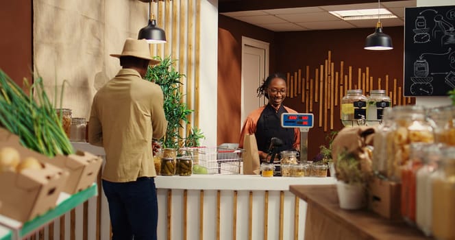 African american seller working at supermarket cash register counter, weighting organic additives free fruits and vegetables. Vendor selling fresh bio produce to male customer at checkout. Camera 2.