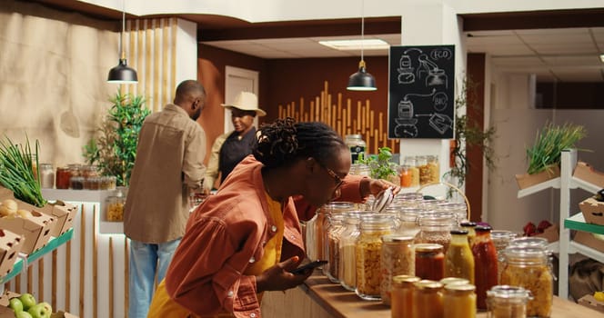 Customer checking shopping list on phone before choosing products, trying to buy organic locally grown food for low carbon footprint. African american woman looking at checklist notes. Camera 2.