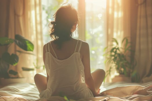 A woman is sitting on a bed with her back to the camera. She is wearing a white robe and she is relaxed. The room has large windows that let in natural light